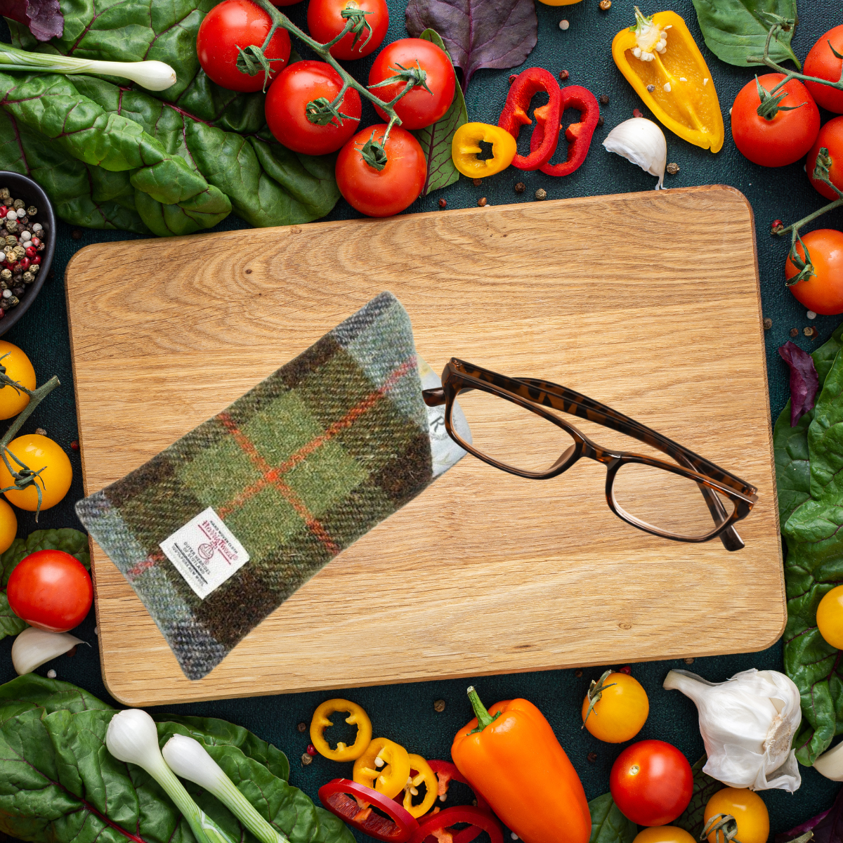 Wooden cutting board with glasses and a woven pouch surrounded by fresh vegetables on a dark surface